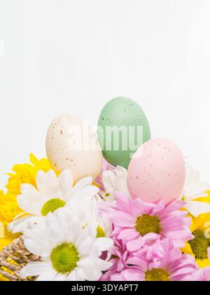 Close-up view of three colorful speckled Easter eggs resting among blooming white, pink, and yellow daisies against a bright white background. Stock Photo