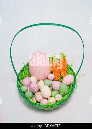 Green wicker basket filled with colorful speckled Easter eggs and decorative carrots on a white background. Stock Photo