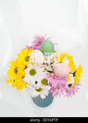A high-angle view captures a vibrant spring bouquet of daisies and three decorative speckled Easter eggs arranged in a green vase on a white surface. Stock Photo