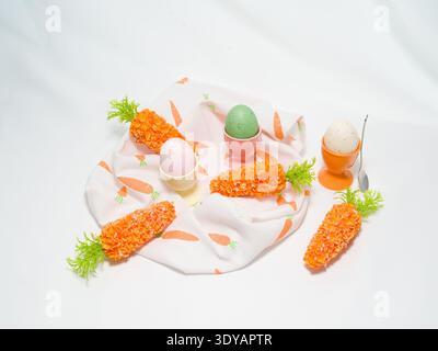 Three colorful speckled Easter eggs in pastel ceramic cups with decorative carrots and a spoon arranged on a folded patterned napkin. Stock Photo