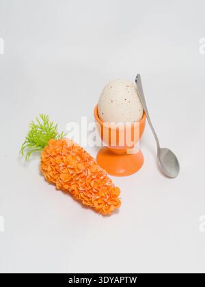 Minimalist Easter composition featuring one speckled egg in an orange ceramic cup with a decorative carrot and a metal spoon on a white background. Stock Photo