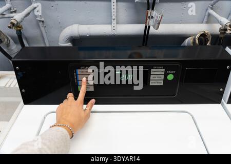 Close-up of a woman’s hand pressing the start button on a washing machine in a laundromat in the United States. Everyday laundry routine Stock Photo