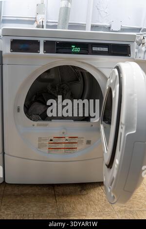 Clothes dryer in a laundromat with the door open, showing the interior drum. A white laundry basket stands nearby. Coin-operated self-service laundry Stock Photo