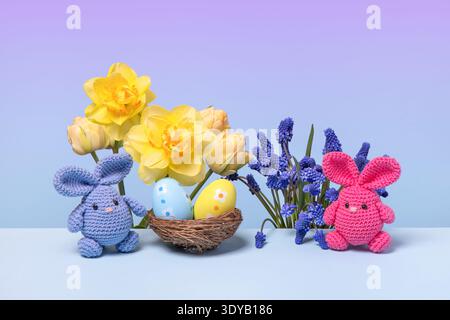 Pink and blue Easter knitted bunnies with multicolored eggs and spring muscari flowers, tulips and daffodils on the blue podium. Concept of Easter Stock Photo
