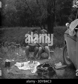 Mikhail Ilyich Kravelis and a close male friend sit comfortably on small folding camp chairs by a picnic blanket, deeply engaged in a friendly conversation in the Donetsk region during the spring of the 1950s. The two young Soviet men relax in the picturesque wild nature right beside their parked classic car. This candid black-and-white photograph beautifully captures the essence of male friendship, relaxed weekend auto tourism, and the wonderful everyday life of the peaceful Donbass before the war. Stock Photo