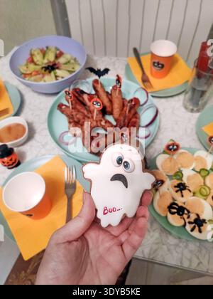 Hand holding a ghost-shaped Halloween cookie with the word “Boo,” surrounded by festive snacks, orange cups, sausages, deviled eggs, and decorations o Stock Photo