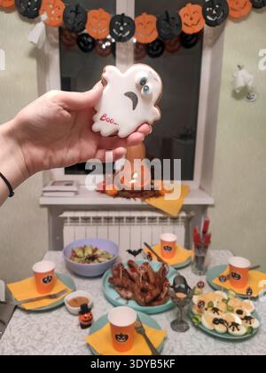 Hand holding a ghost-shaped Halloween cookie with the word “Boo,” surrounded by festive snacks, orange cups, sausages, deviled eggs, and decorations o Stock Photo