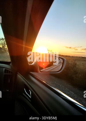 View from inside a car of the setting sun reflected in the side mirror on a country road, creating a warm atmosphere and a sun glare effect. Stock Photo