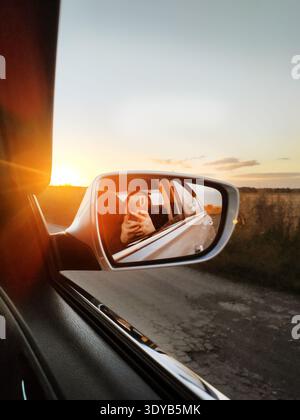 View from inside a car of the setting sun reflected in the side mirror on a country road, creating a warm atmosphere and a sun glare effect. Stock Photo