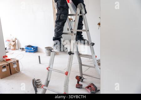 Worker carefully standing on a portable aluminum ladder, wearing safety work boots and pants, performing renovation tasks in a residential or commerci Stock Photo