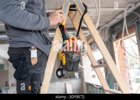 Construction worker installing wiring on ladder with tools Stock Photo