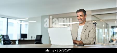 Happy mature bank manager looking at laptop in office sitting at desk. Stock Photo