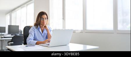 Busy elegant mid aged business woman using laptop working in corporate office. Stock Photo
