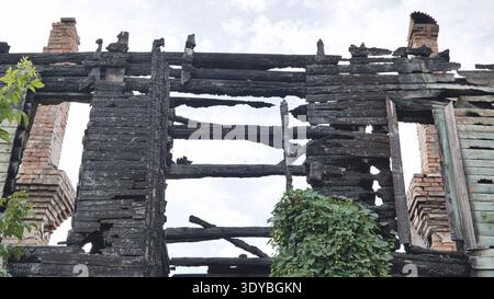 Charred remains of a burnt wooden house with brick chimneys against a cloudy sky Stock Photo