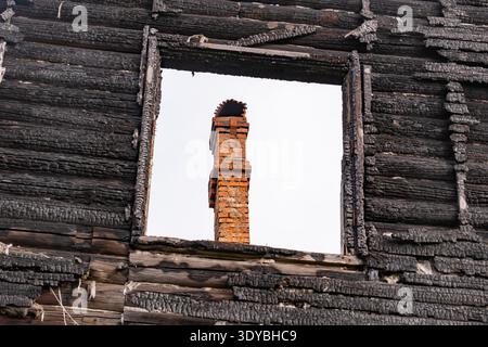 Charred log house wall with brick chimney seen through a window frame Stock Photo