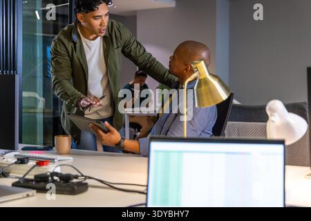 Two male coworkers in office, wearing blazer and overshirt, discussing over tablet and laptop Stock Photo