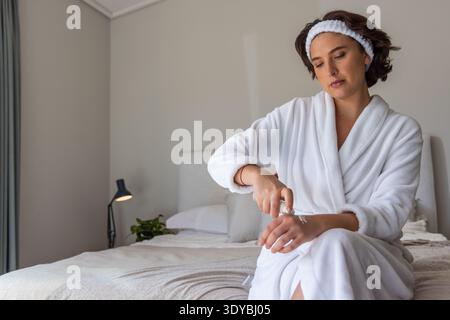 Woman in white bathrobe and headband sitting on bed applying hand cream from small tube Stock Photo