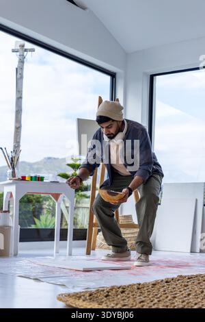 Indian man crouching over floor canvas in art studio, holding wooden mixing bowl and wearing beanie Stock Photo