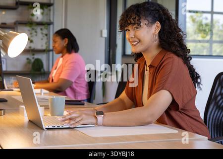 Diverse female coworkers smiling and working at long wooden desk with silver laptop, smartwatch Stock Photo