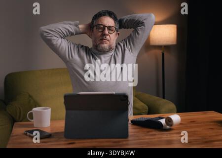 Mature man leaning back with hands behind head after reviewing bills and financial documents at home desk. Concept of financial stress relief, budgeti Stock Photo