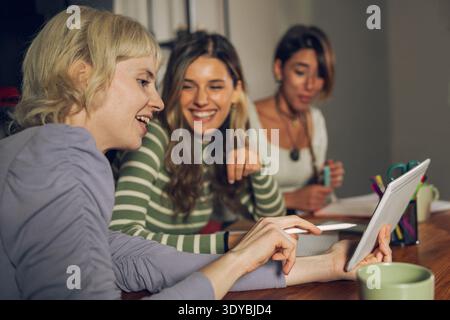 Three young women sitting together at a table while using a digital tablet, smiling and collaborating in an indoor study or creative workspace, modern Stock Photo