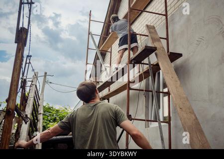 A low-angle shot captures two skilled construction workers diligently engaged in the renovation of a residential building under a partly cloudy sky. O Stock Photo