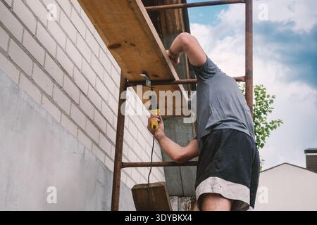 This image captures a dedicated man on scaffolding, meticulously drilling into a brick wall as part of a home renovation project. The focus highlights Stock Photo
