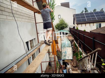 This image captures a real-life scene of two men collaborating on a home improvement project. One man stands on scaffolding, guiding a long wooden pla Stock Photo