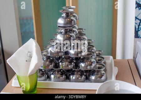 Pyramid stack of polished metal ice cream cups on white plastic tray in cafe interior, green glass with white napkins in foreground, professional kitc Stock Photo