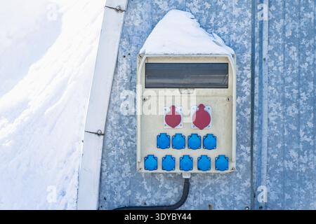 Industrial electrical outlet panel mounted outside with snow cap, blue plug sockets and red power connectors on metal siding, temporary power distribu Stock Photo