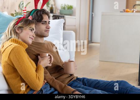 Couple wearing antler candy cane headbands sitting on white couch in living room holding mugs. Festive, cozy, modern, warm, celebration, home decor, joyful || Model released Stock Photo