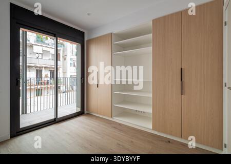 A modern, empty room with wooden flooring and large sliding glass doors. The room features a built-in wardrobe with light wood finish and open shelves Stock Photo
