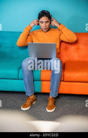 Indian man wearing orange sweater and jeans sitting on two-tone sofa placing headset, using laptop. Remote, workstation, studio, casual, focused, colorful, technology || Model released Stock Photo