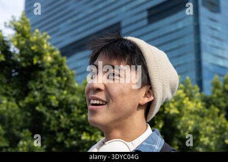 Asian teenage male smiling, looking upward in city park wearing beige beanie and headphones. Urban, greenspace, outdoors, denim, backpack, glass, modern || Model released Stock Photo