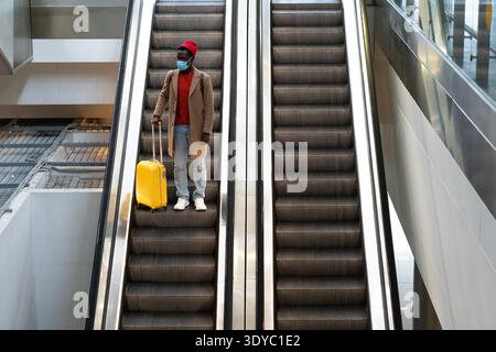 Top view of Black man with yellow luggage standing on escalator at airport terminal wearing face mask during flu season. || Model released Stock Photo