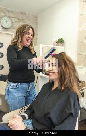 Professional hairstylist straightening a smiling woman's hair with a flat iron in a bright busy salon, creating a polished new look during a friendly Stock Photo