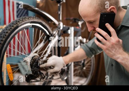 Bearded young man repairing broken bicycle at home and calling mechanic on smartphone for professional advice. Copy space Stock Photo