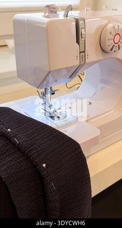 Sewing machine with black fabric and pins positioned on a white table, showcasing the needle and presser foot in a well-lit indoor workspace Stock Photo