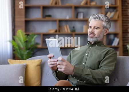 Man with grey hair and beard sitting comfortably on a sofa, browsing information on a digital tablet, connecting with technology in a cozy domestic environment Stock Photo