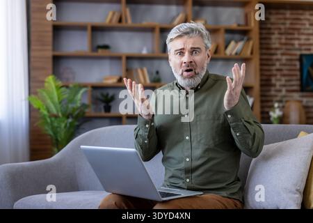 Senior man sitting on a sofa, looking surprised and frustrated at his laptop, experiencing confusion or a technical problem while using online technology at home Stock Photo