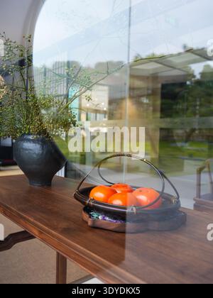 Orange persimmons in a traditional woven basket and a dark vase with green branches sitting on a wooden table, viewed through a circular glass window. Stock Photo