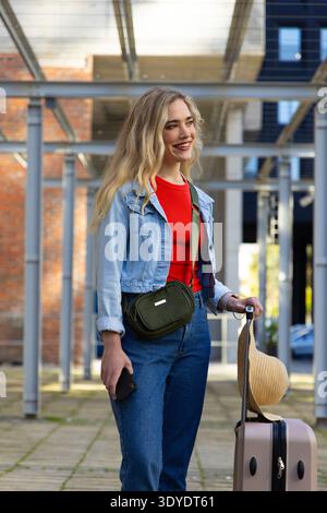 Adult woman standing smiling holding suitcase handle and checking smartphone under metal pergola Stock Photo