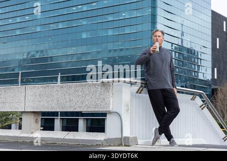 Middle-aged man in gray pullover leaning on handrail sipping bottle on overpass, copy space Stock Photo