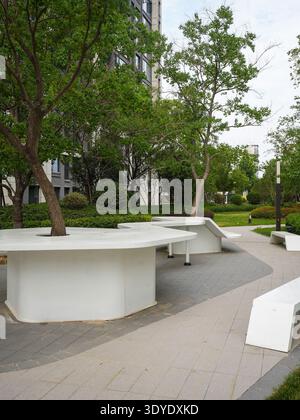 A minimalist, white sculptural garden bench set in a quiet residential courtyard with tall, leafy trees. The image conveys architectural elegance, med Stock Photo