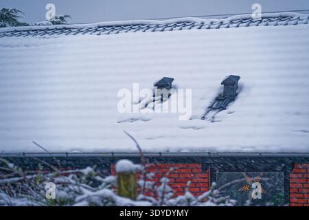 A house roof covered in fresh snow with two chimneys and falling snowflakes during winter. The brick wall and window with star decoration are visible. Stock Photo