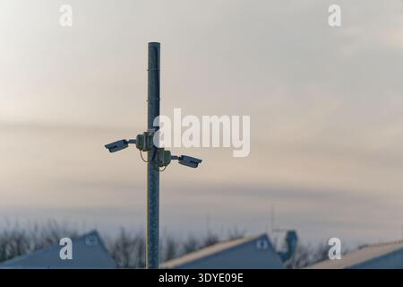 Two security cameras attached to a tall metal pole monitoring an outdoor area with buildings and trees in the distance under a cloudy sky. Stock Photo