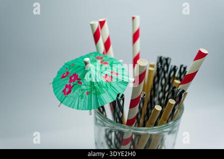 A variety of colorful paper straws with red white black and natural tones along with a green floral cocktail umbrella in a clear glass container. Stock Photo
