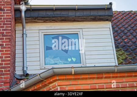 A damaged window with shattered glass on the upper floor of a house featuring brick walls, tiled roof, and metal gutters under clear sky conditions. Stock Photo