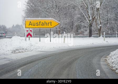 Winter scene showing a snow covered road curving beside a yellow exit sign with snow on trees and ground in a cold forest environment. Stock Photo