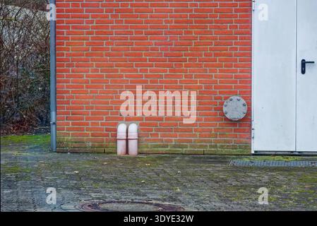 Red brick wall featuring two white pipes and a white door with a black handle on a paved ground covered with moss and fallen leaves outdoors. Stock Photo